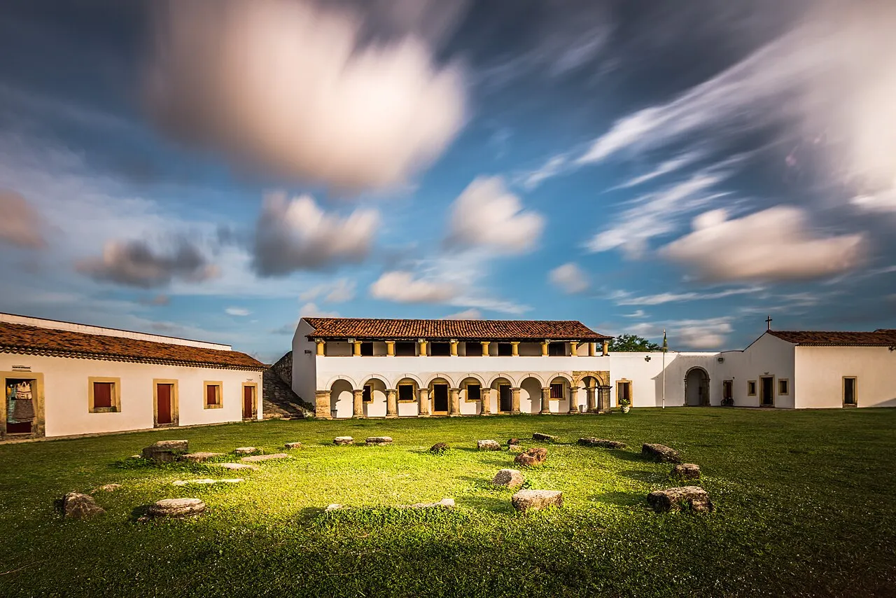 O pátio gramado da Fortaleza de Santa Catarina, em Cabedelo, com as construções brancas ao fundo e nuvens em movimento.