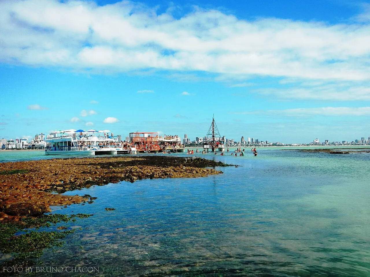 O recife de Picãozinho exposto na maré baixa, com os barcos de passeio ancorados e os prédios da orla ao fundo.