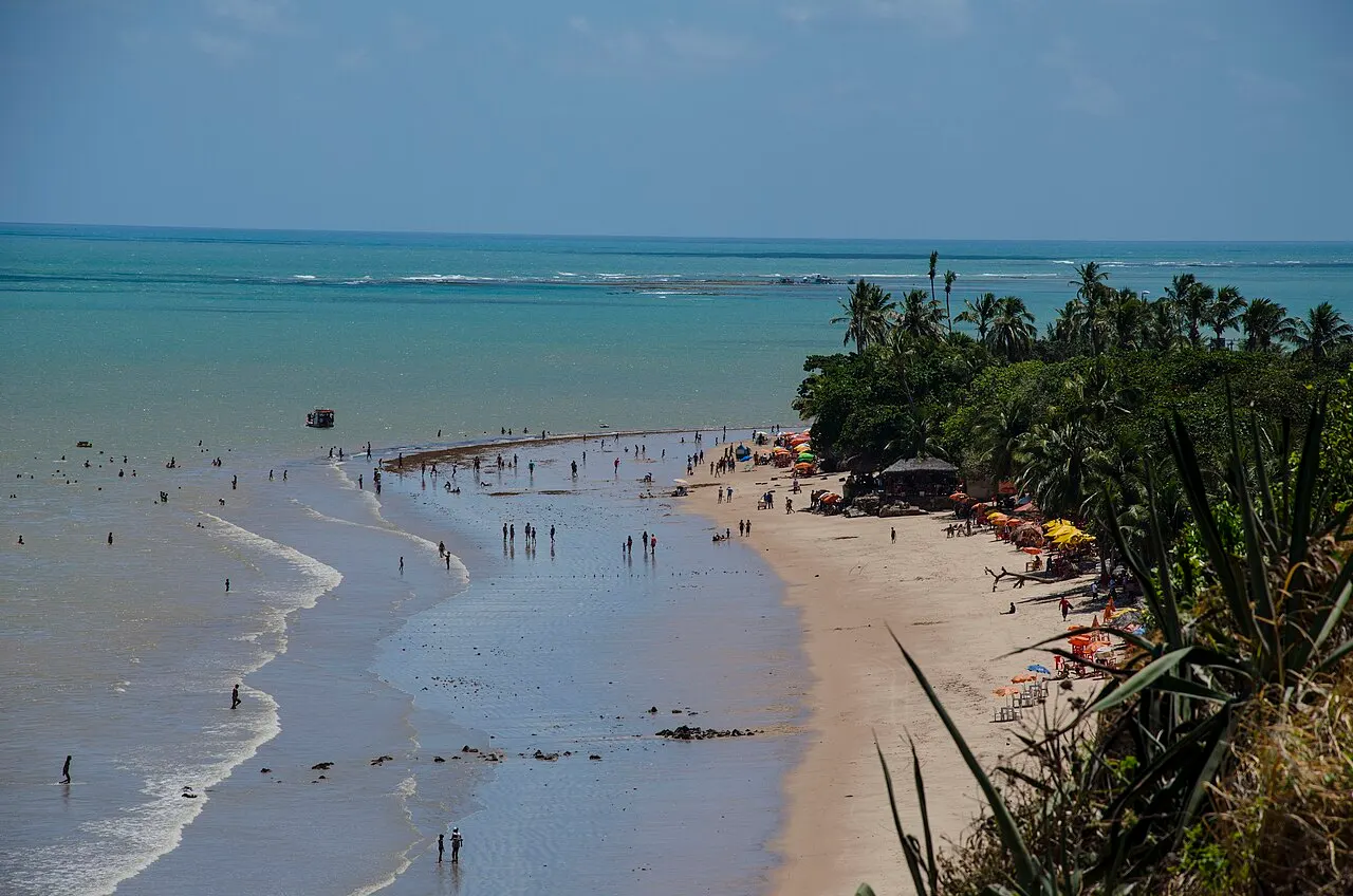 A praia da Ponta do Seixas vista do alto da falésia, com a maré baixa e gente caminhando na água rasa.