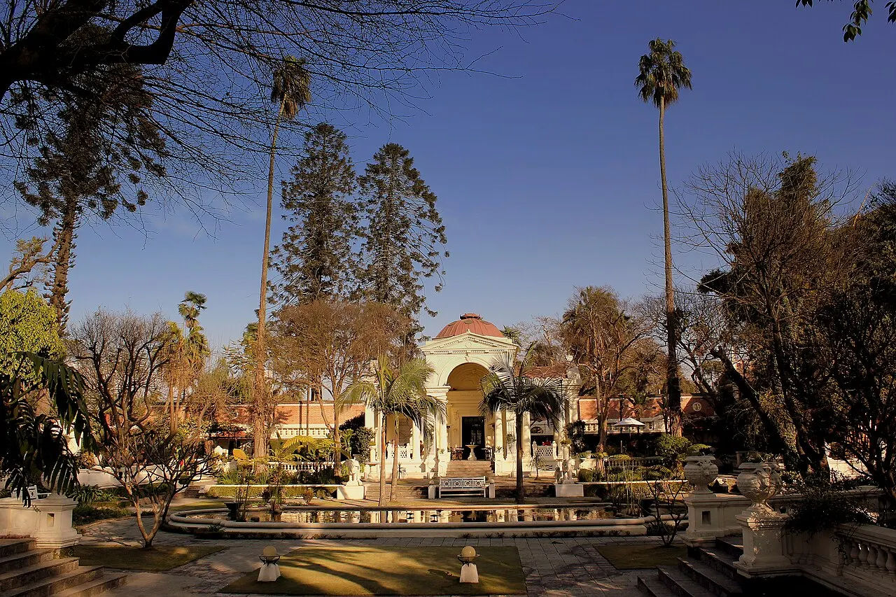Pavilhão branco e rosado com uma cúpula central e colunas, entre árvores e palmeiras.