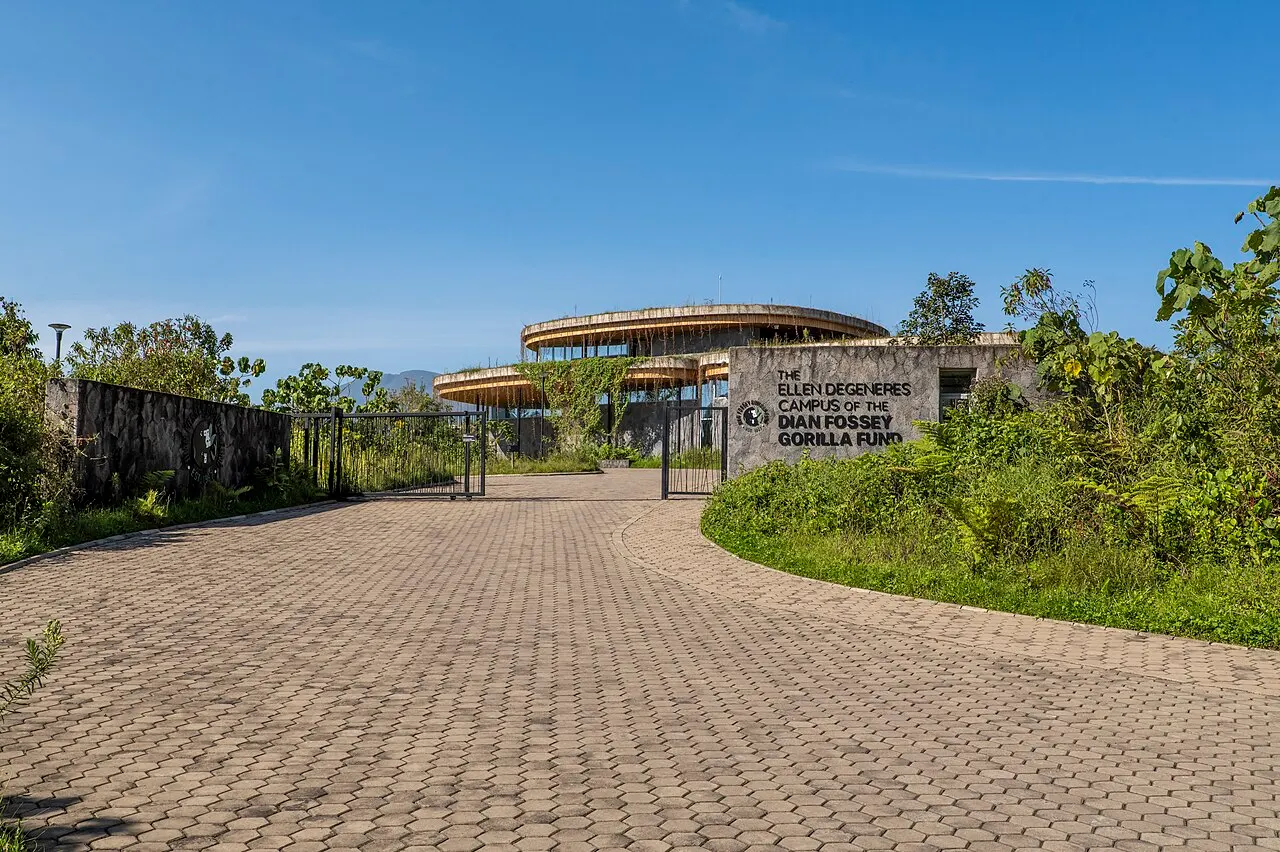 A paved driveway leading to a gate, with a stone wall bearing the campus name and, behind it, multi-level circular buildings among the vegetation, under a blue sky.