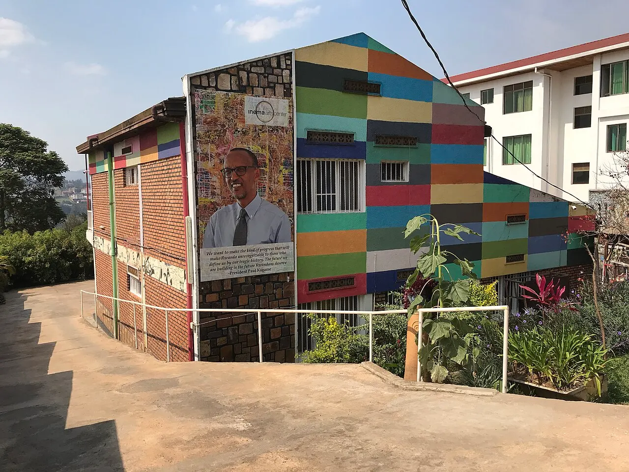Brick building with its façade painted in rectangular bands of many colours and a large panel with a painted portrait, beside a concrete ramp and a garden.