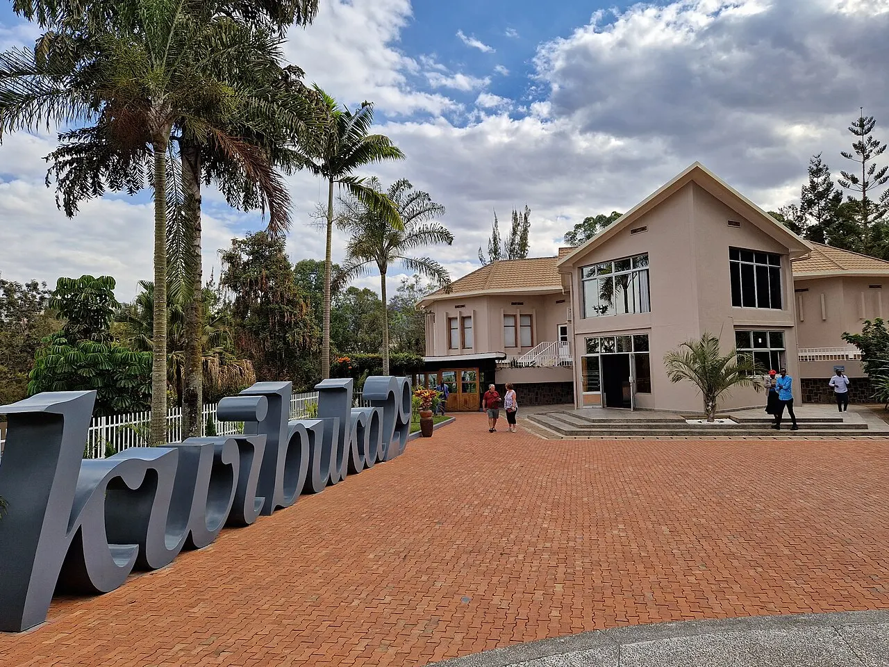 A two-storey pinkish building surrounded by palm trees, with a red-brick courtyard in front, large grey metal letters on the left and a few small figures near the entrance.