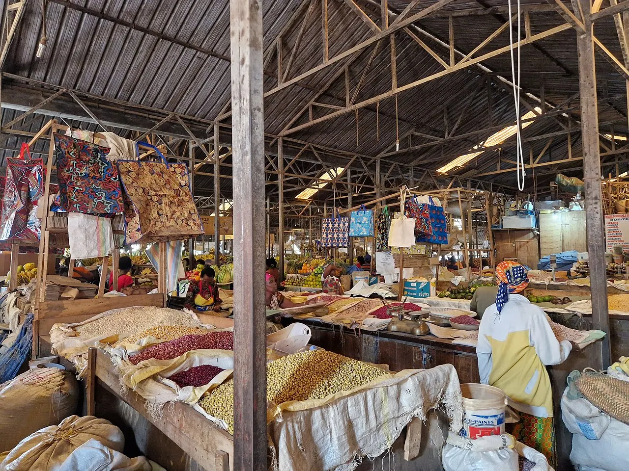 Inside a market under a metal roof on a wooden frame, with stalls of beans and grains in open sacks, patterned fabrics hanging and a vendor wearing a headscarf.