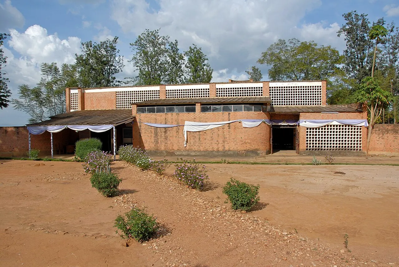 A low brick church with white and purple fabric draped along the façade and openwork block walls at the top, in front of a red-earth yard with small shrubs.