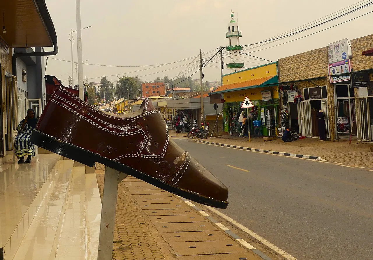 Sculpture of a giant brown shoe on a post on the pavement, in front of a paved street lined with colourful shops and a green and white minaret in the background.