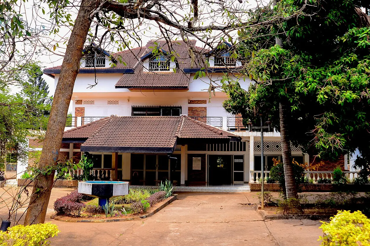 A large house with white walls and brown roofs and balconies upstairs, seen between trees, with a flower bed and a small fountain in front of the entrance.