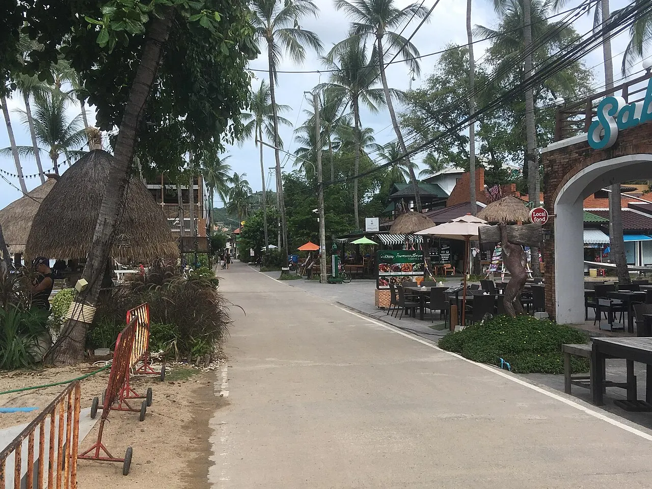 Une rue étroite et vide à Bophut, avec des tables de restaurant et des parasols d'un côté, un kiosque en paille et des cocotiers de l'autre.