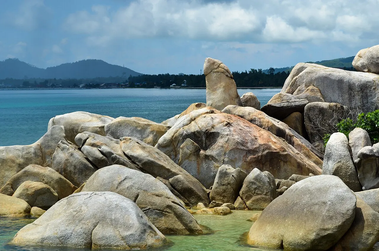 De gros rochers de granit arrondis au bord d'une mer bleu-vert, avec une pierre haute et étroite dressée au centre et des collines au loin.
