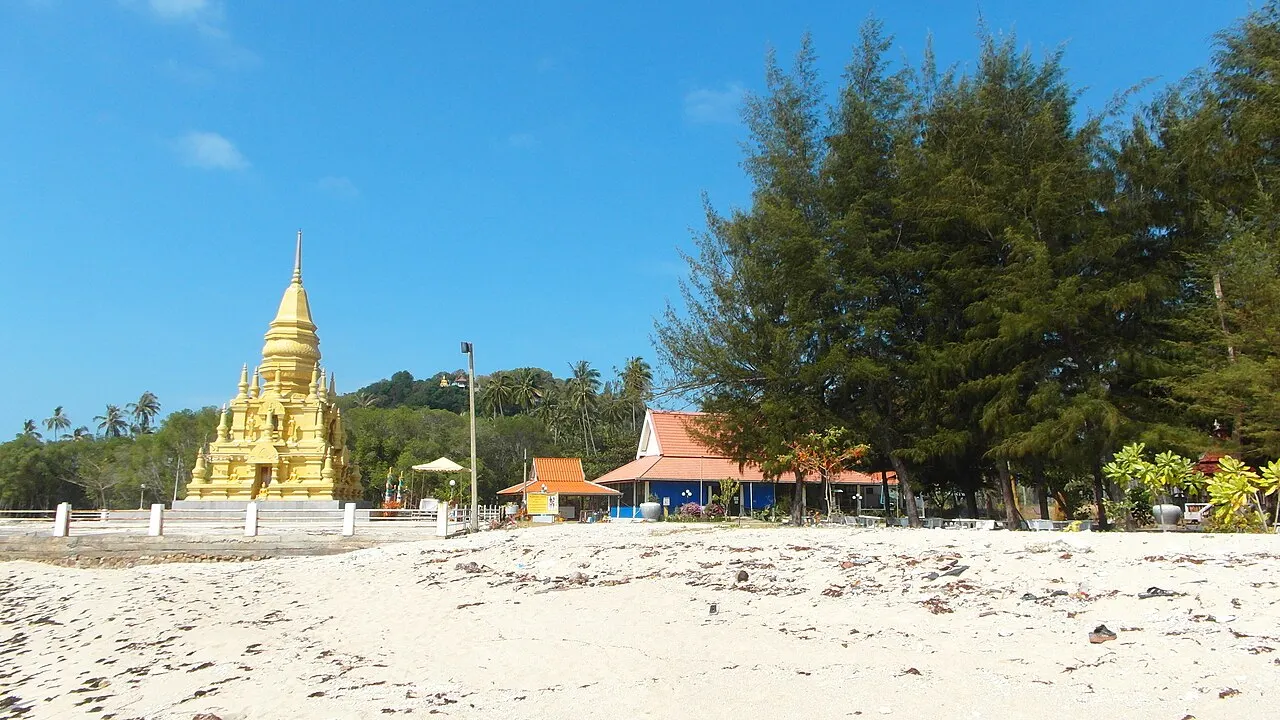 Un chedi jaune et pointu au bord d'une plage de sable clair, avec des filaos et des bâtiments aux toits orange à côté, sous un ciel bleu.