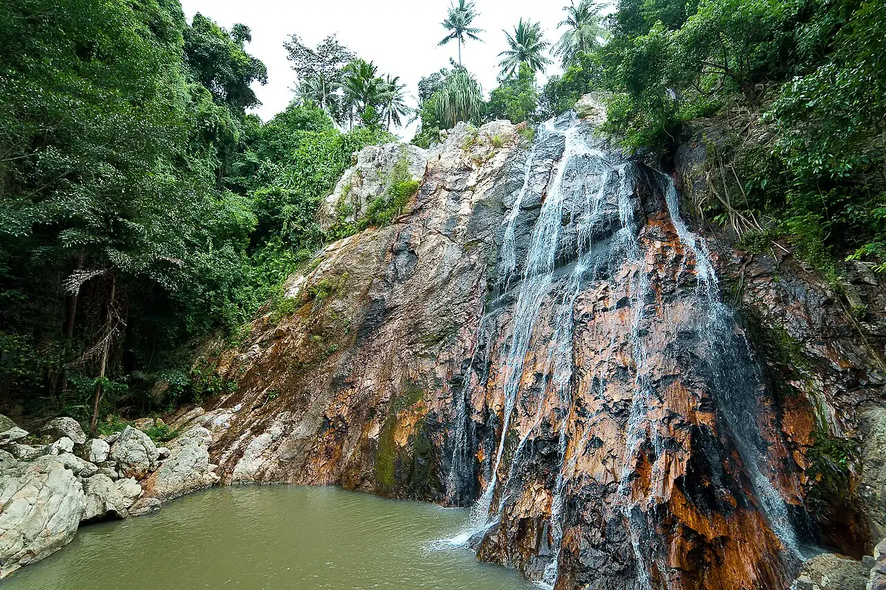 Une cascade qui dévale une paroi de roche orangée et sombre jusqu'à un bassin d'eau verdâtre, entourée de forêt et de cocotiers.