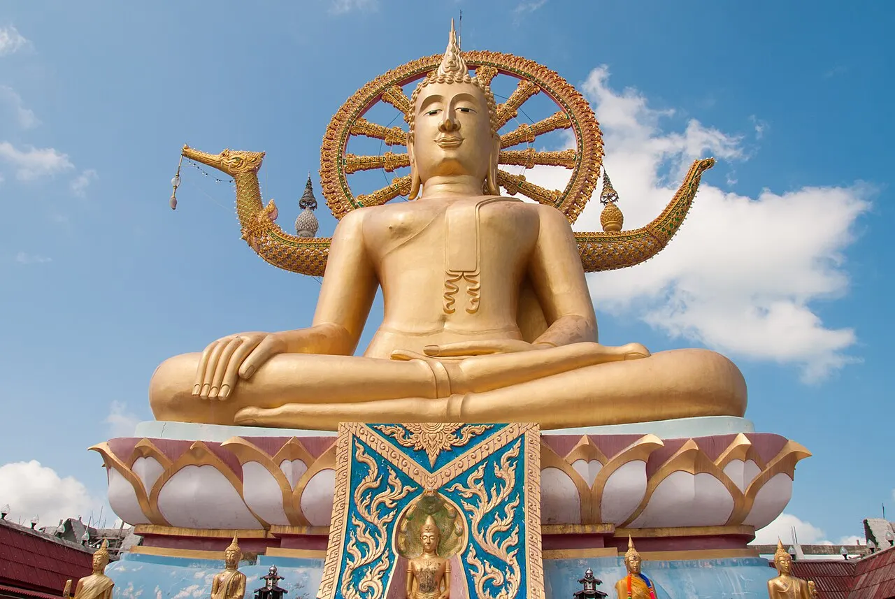 La grande statue dorée du Bouddha assis, avec une auréole en forme de roue derrière la tête, sur un piédestal en pétales de lotus, devant un ciel bleu nuageux.