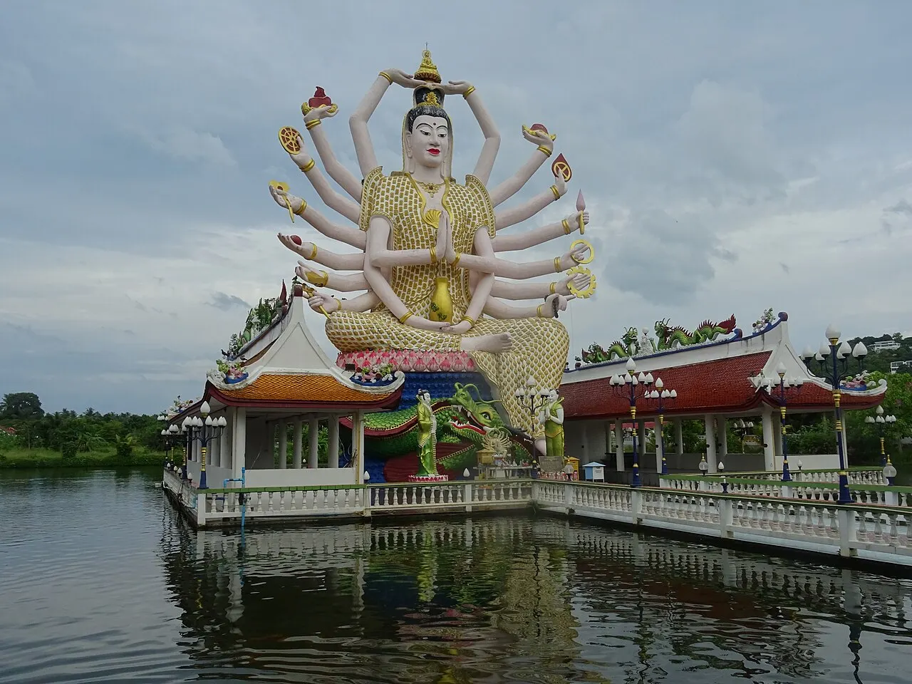 La statue blanche et dorée de Guanyin, aux nombreux bras déployés en éventail, sur un pavillon au milieu d'un lac, reflétée dans l'eau sous un ciel nuageux.