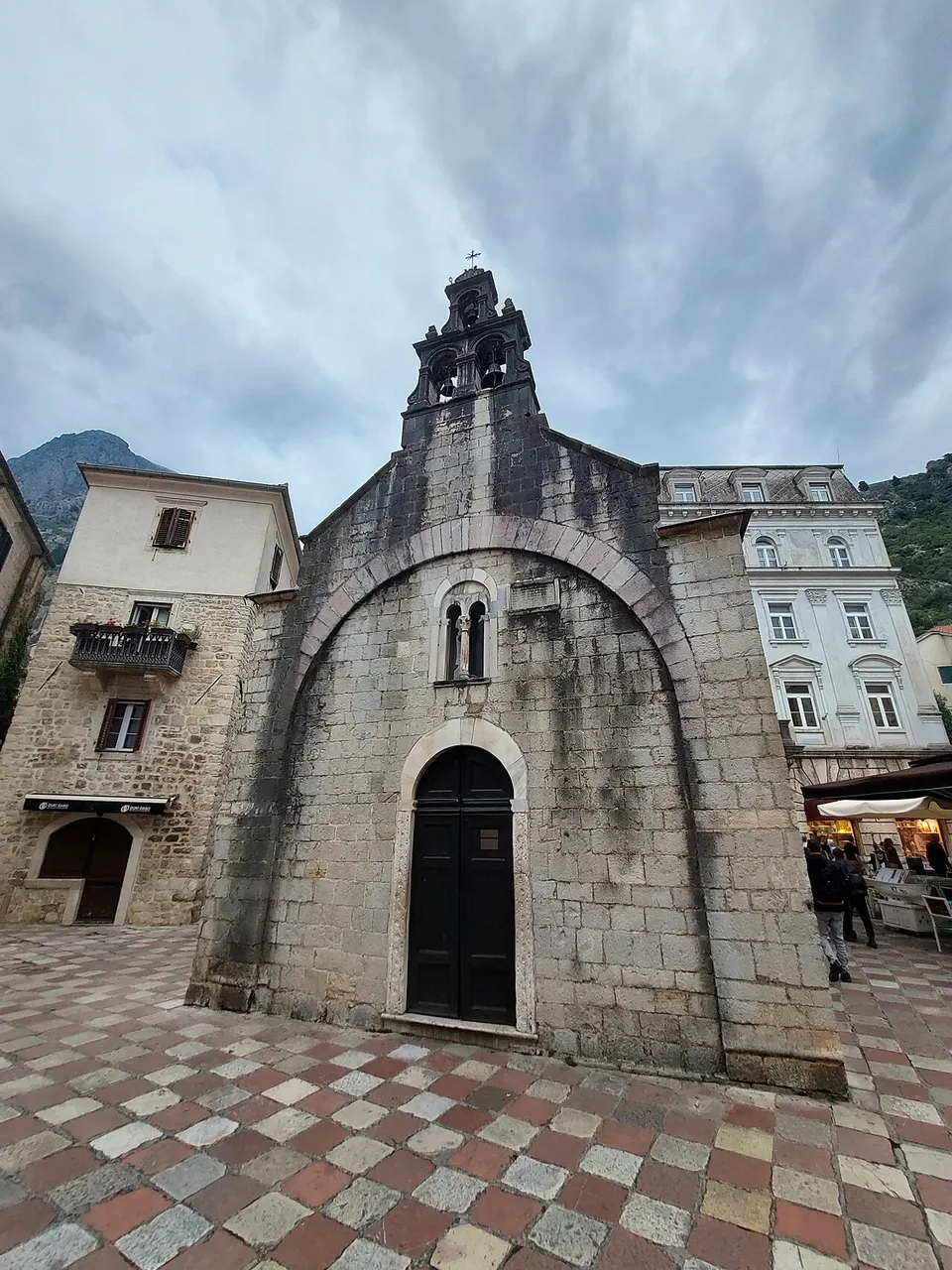 A pequena Igreja de São Lucas, de pedra cinzenta e campanário de parede, numa praça de piso xadrez entre prédios claros.
