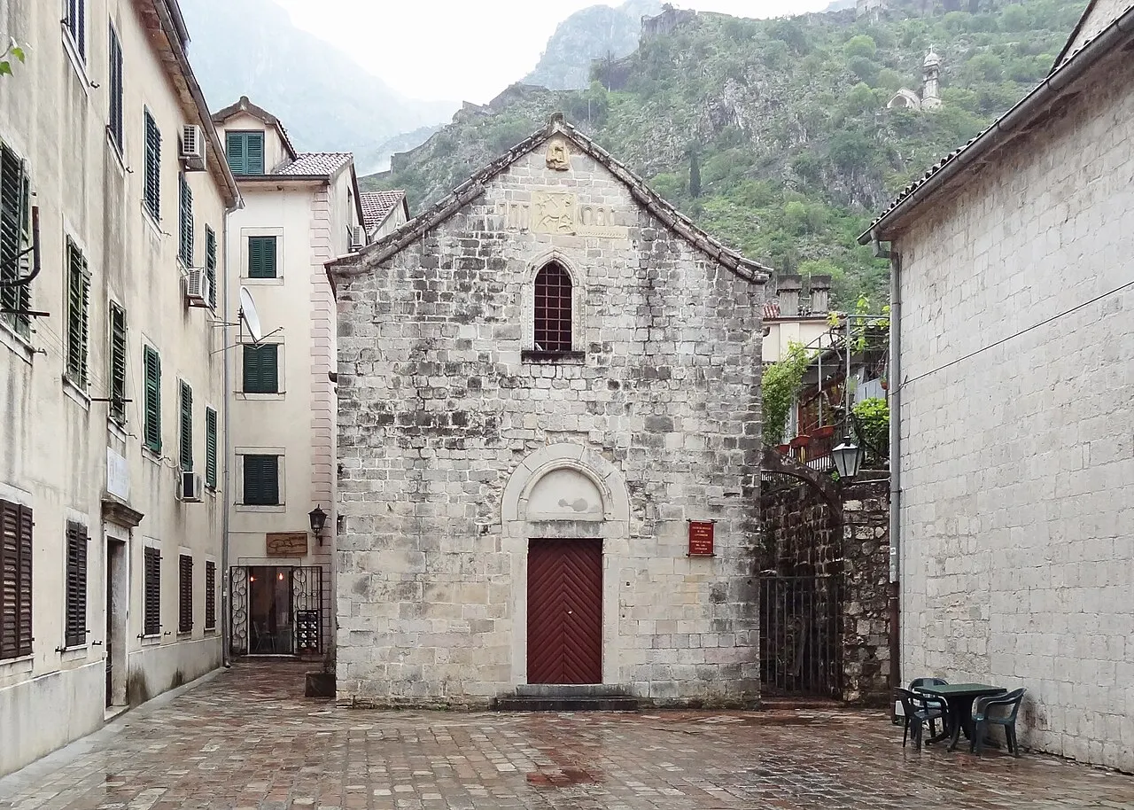 A fachada de pedra clara da Igreja de São Miguel, com uma janela estreita e porta vermelha, numa pracinha molhada entre prédios.