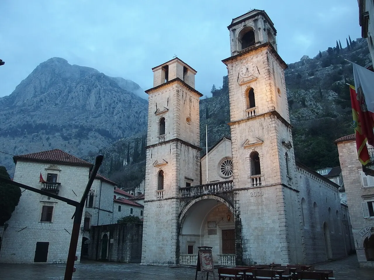 A fachada de pedra da Catedral de São Trifão, com duas torres, a rosácea e o arco da entrada, ao entardecer, com a montanha atrás.