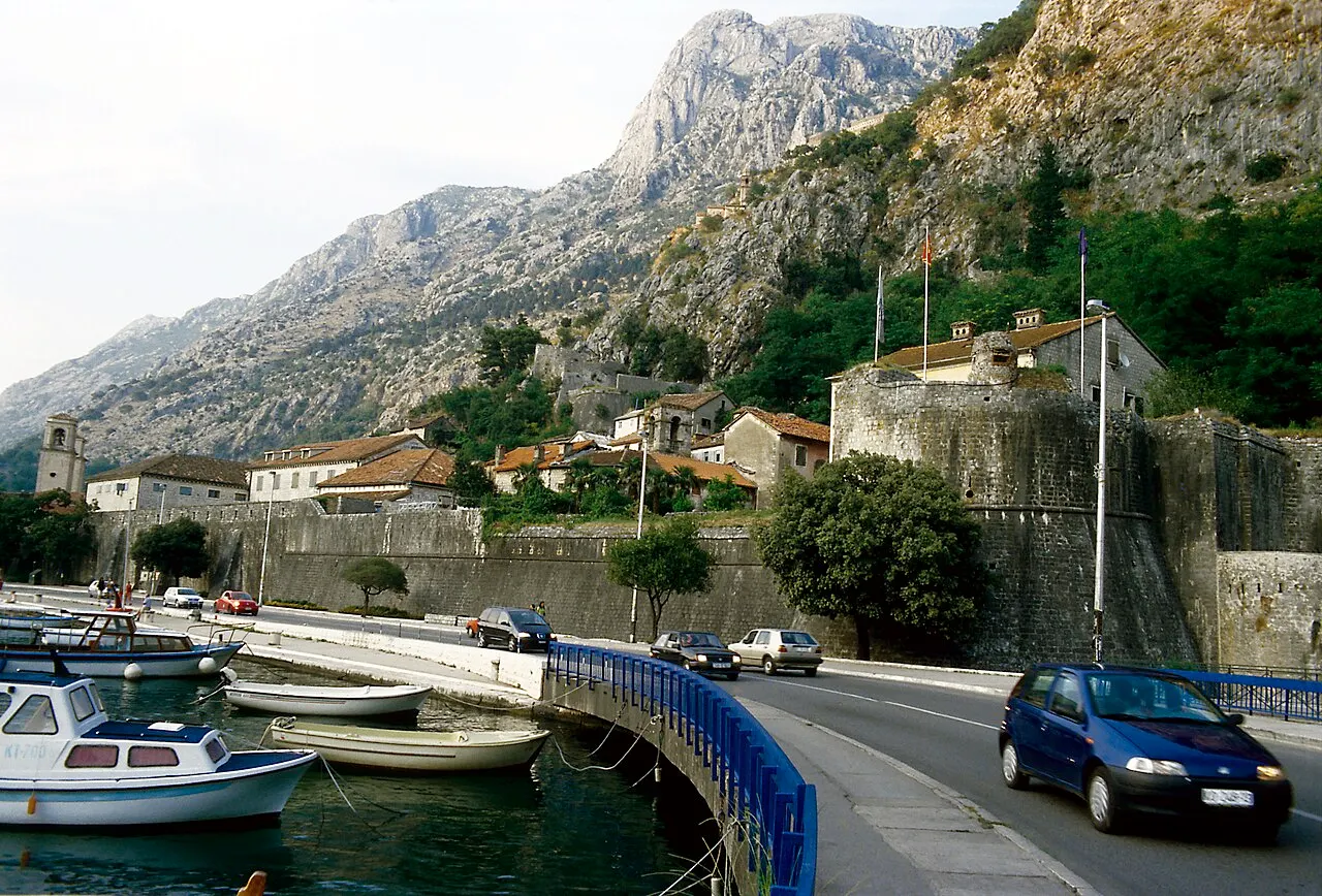 A muralha de pedra de Kotor junto ao fosso com barcos, uma ponte com carros e as fortificações subindo a montanha.