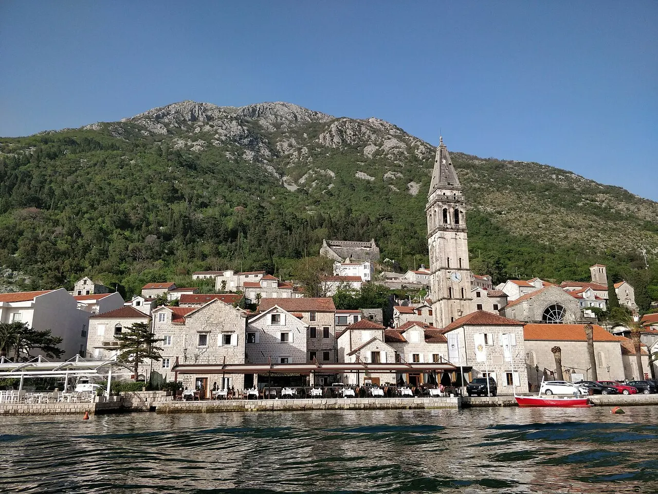 Perast vista de um barco: casas de pedra na beira da água, o campanário alto de São Nicolau e o morro verde atrás.
