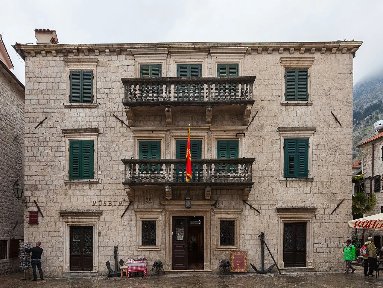 A fachada de pedra do palácio do Museu Marítimo, com persianas verdes, duas sacadas de balaústres e a bandeira de Montenegro.