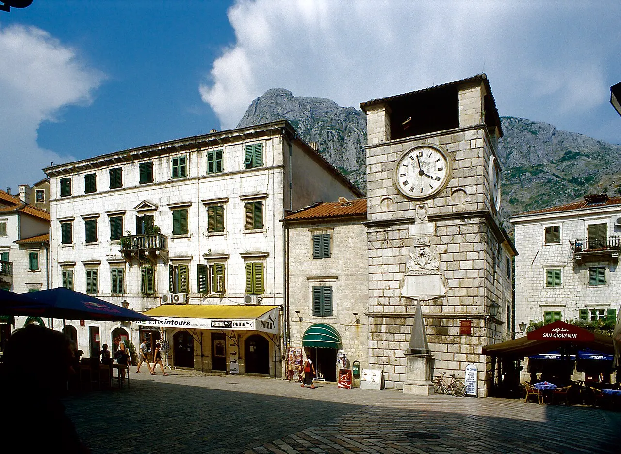 A Praça das Armas de Kotor, com a Torre do Relógio, prédios claros de persianas verdes e a montanha rochosa atrás.