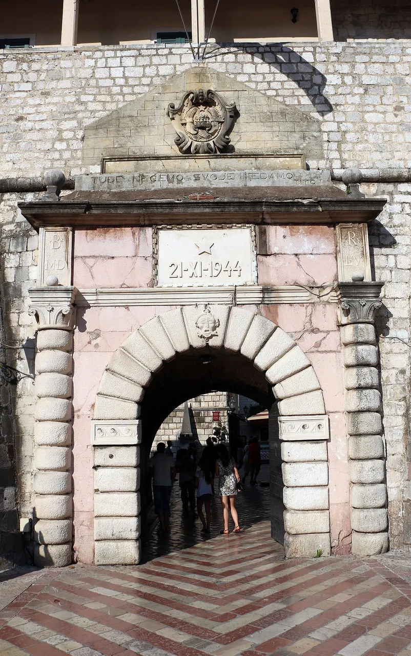 A Porta do Mar de Kotor, um arco de pedra com moldura rosada, a data 21-XI-1944 acima e pessoas passando pela passagem.