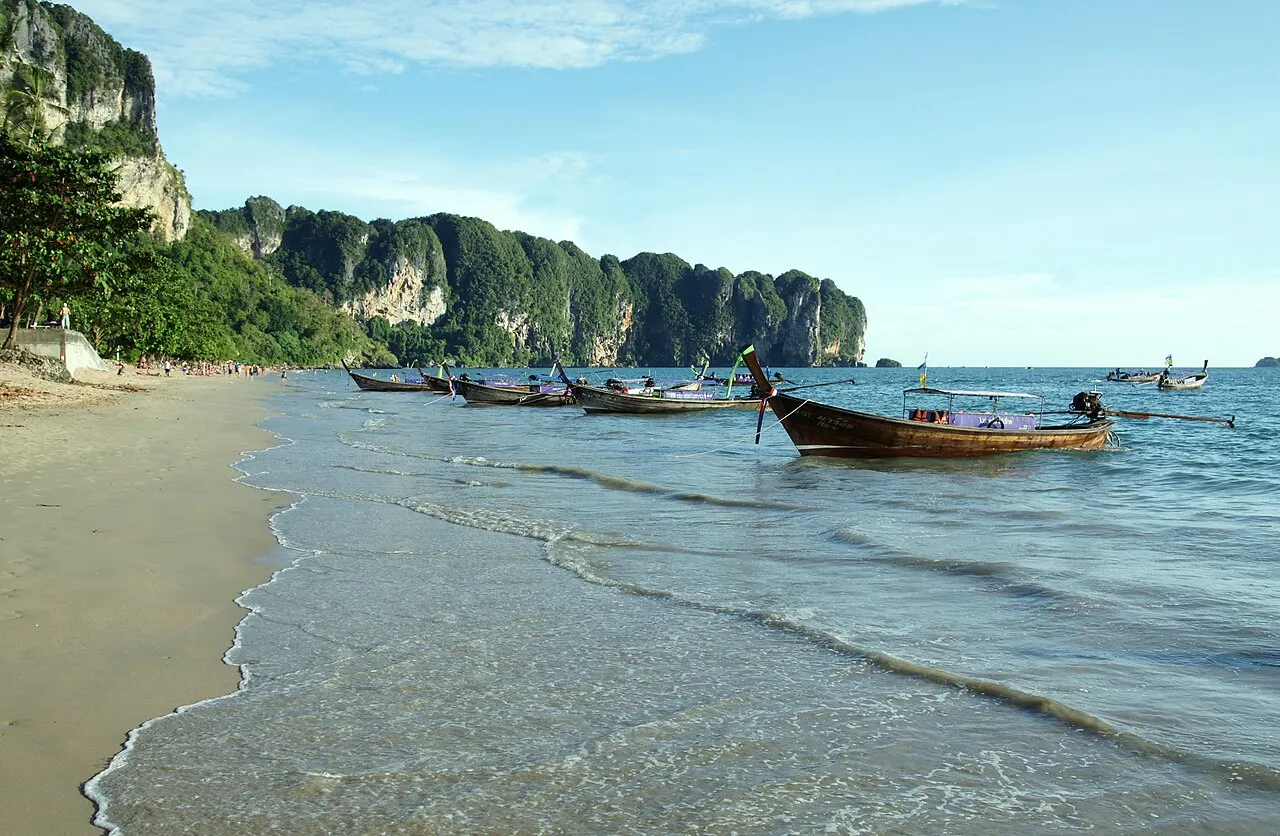 Bateaux à longue queue au mouillage au bord de la plage d'Ao Nang, avec le sable à gauche et des collines calcaires boisées en arrière-plan.