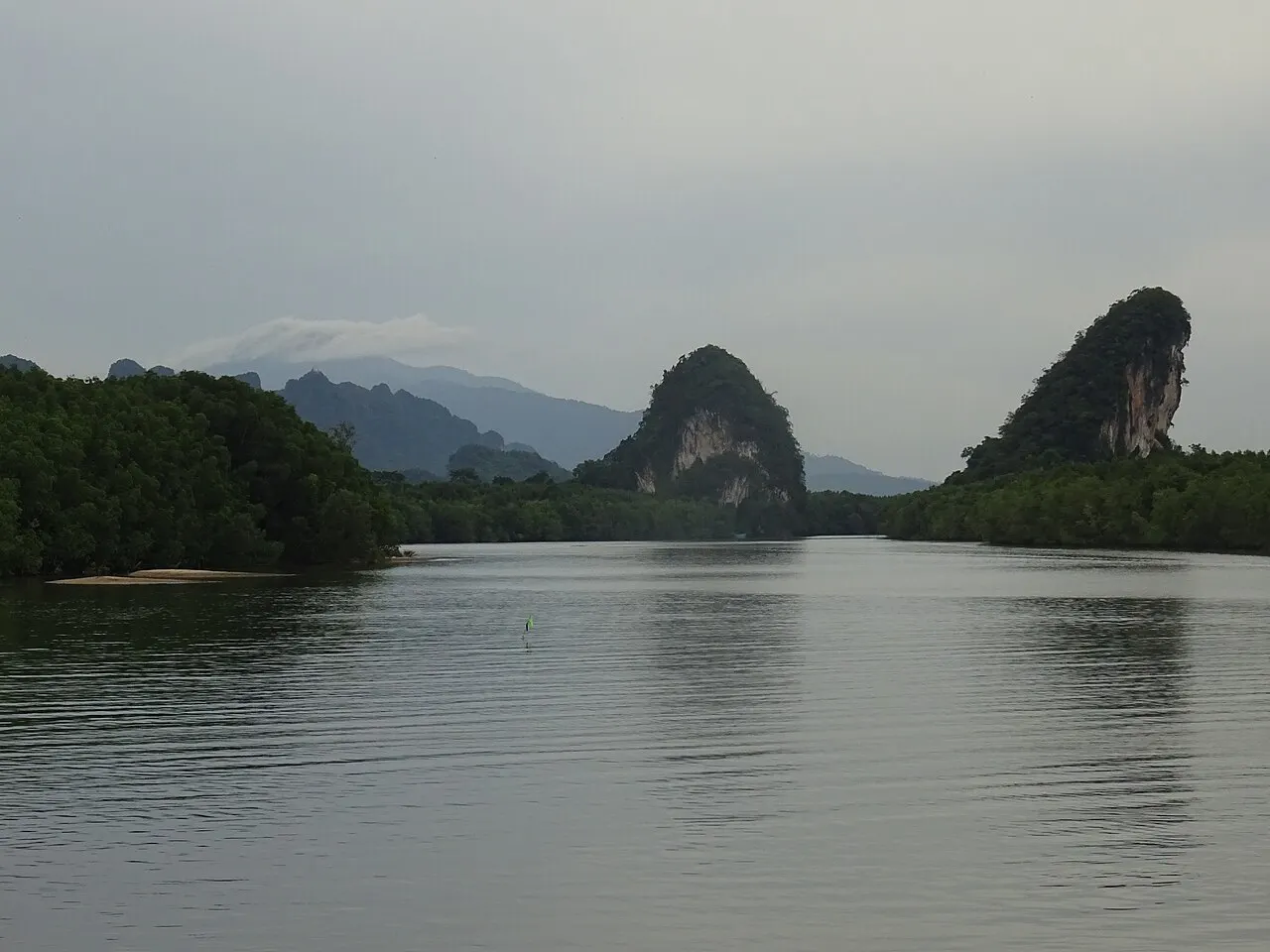 Deux collines calcaires dressées au-dessus de la mangrove, de part et d'autre de la rivière Krabi, avec l'eau calme au premier plan et des montagnes au fond par temps couvert.
