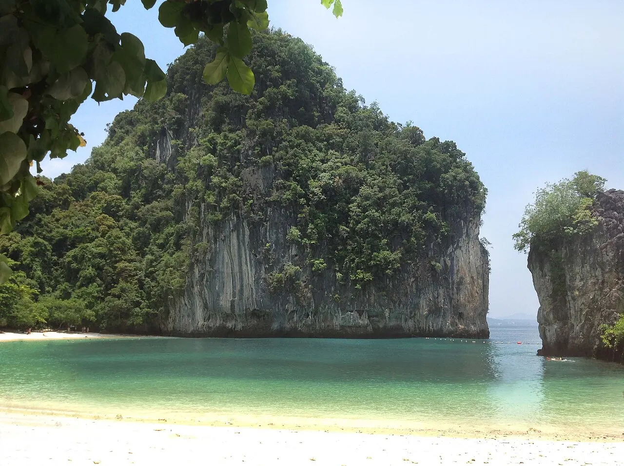 Une crique de sable blanc et d'eau vert clair à Ko Hong, fermée par des parois calcaires couvertes de forêt, avec des feuilles au premier plan en haut.