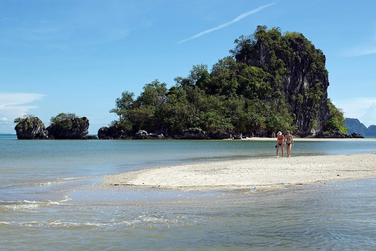 Un banc de sable clair dans l'eau peu profonde de la plage de Noppharat Thara, avec deux personnes marchant au loin et un îlot calcaire couvert d'arbres.