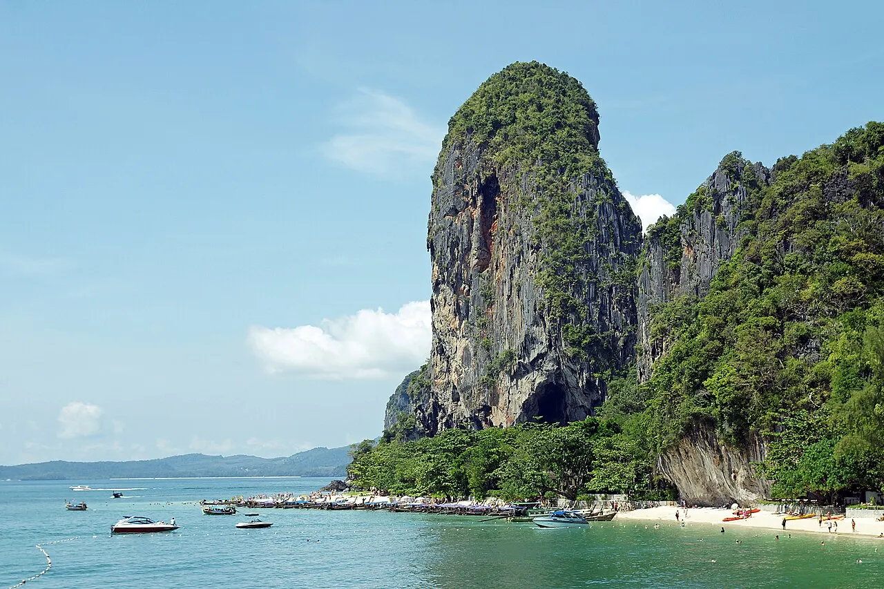 Un haut rocher calcaire percé d'une grotte à sa base tombe sur le sable clair de la plage de Phra Nang, avec des bateaux au mouillage dans l'eau bleu-vert.