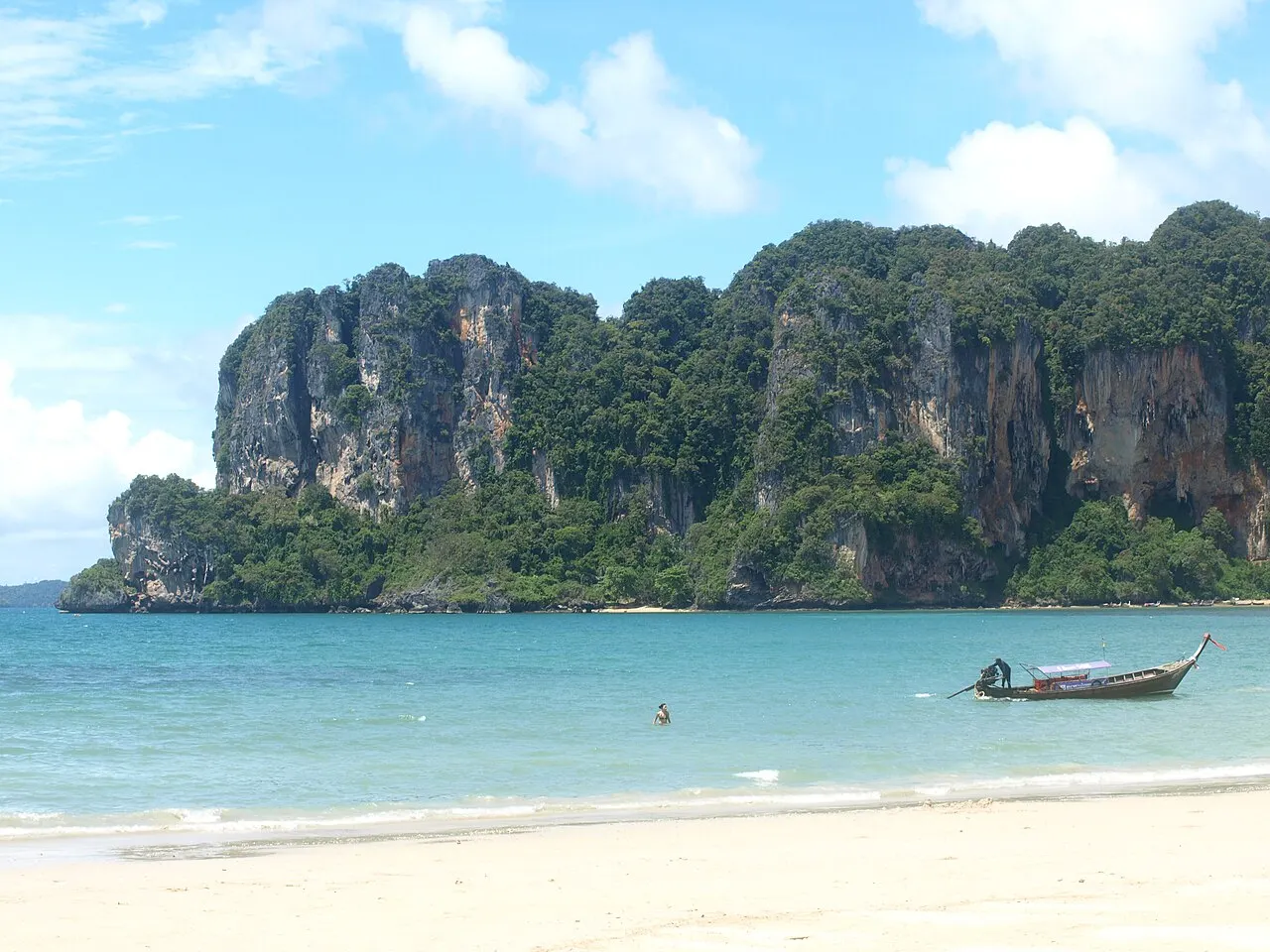 Sable blanc et eau turquoise peu profonde à Railay, avec un bateau à longue queue et un baigneur devant une falaise calcaire couverte de forêt.