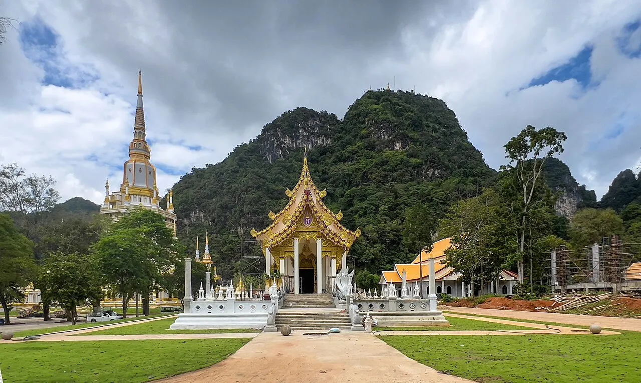 Pavillon blanc et doré et une haute pagode sur la pelouse du Wat Tham Suea, au pied d'une colline calcaire boisée, sous un ciel nuageux.
