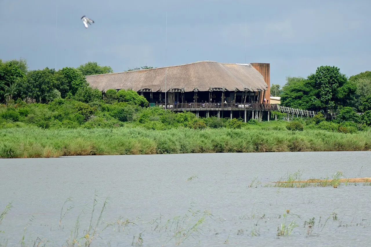 A wide, calm river in the foreground and, on the far bank, a long thatched building with a deck over the bank, among green trees and tall grass.