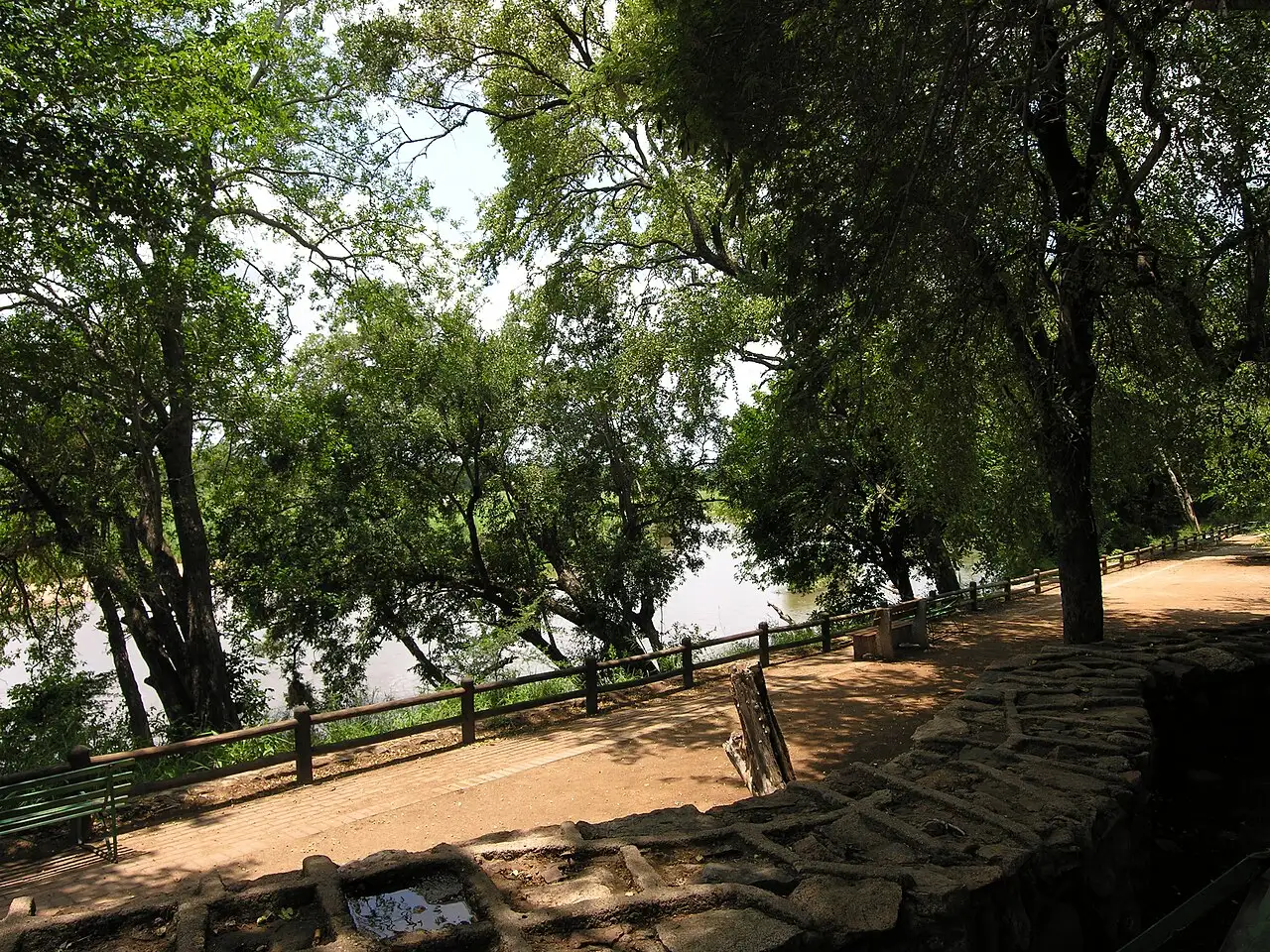Terraza de tierra apisonada con un murete de piedra y una baranda de madera, a la sombra de árboles grandes, con el río asomando entre los troncos al fondo.