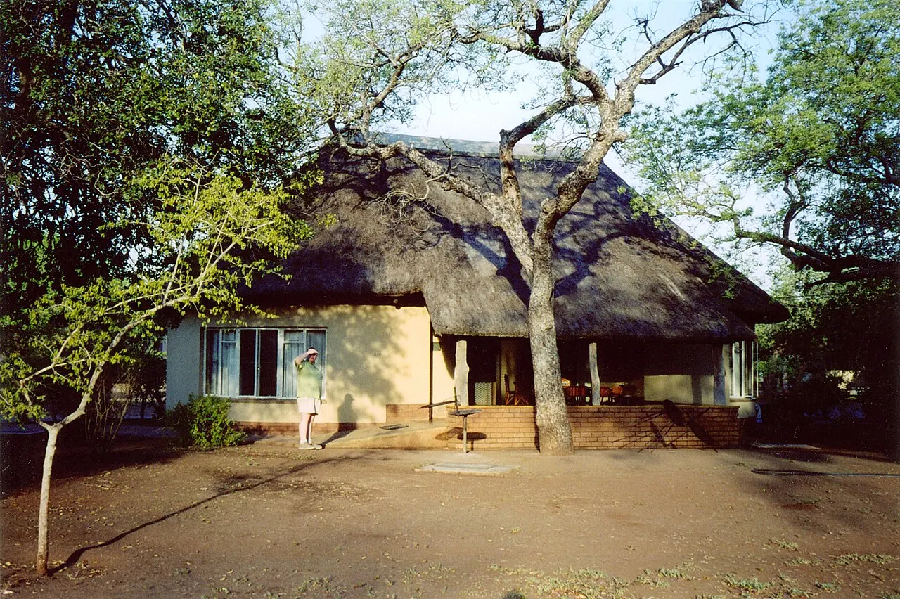 A single-storey cottage with pale walls and a thatched roof, a veranda and a tree in front, bare earth ground and a person standing by the wall shading their eyes from the sun.