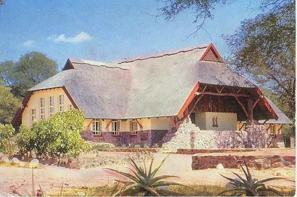 Postal antigua de un edificio de paredes claras con un gran techo de paja, entrada con porche de madera y muro de piedra, rodeado de árboles y aloes.
