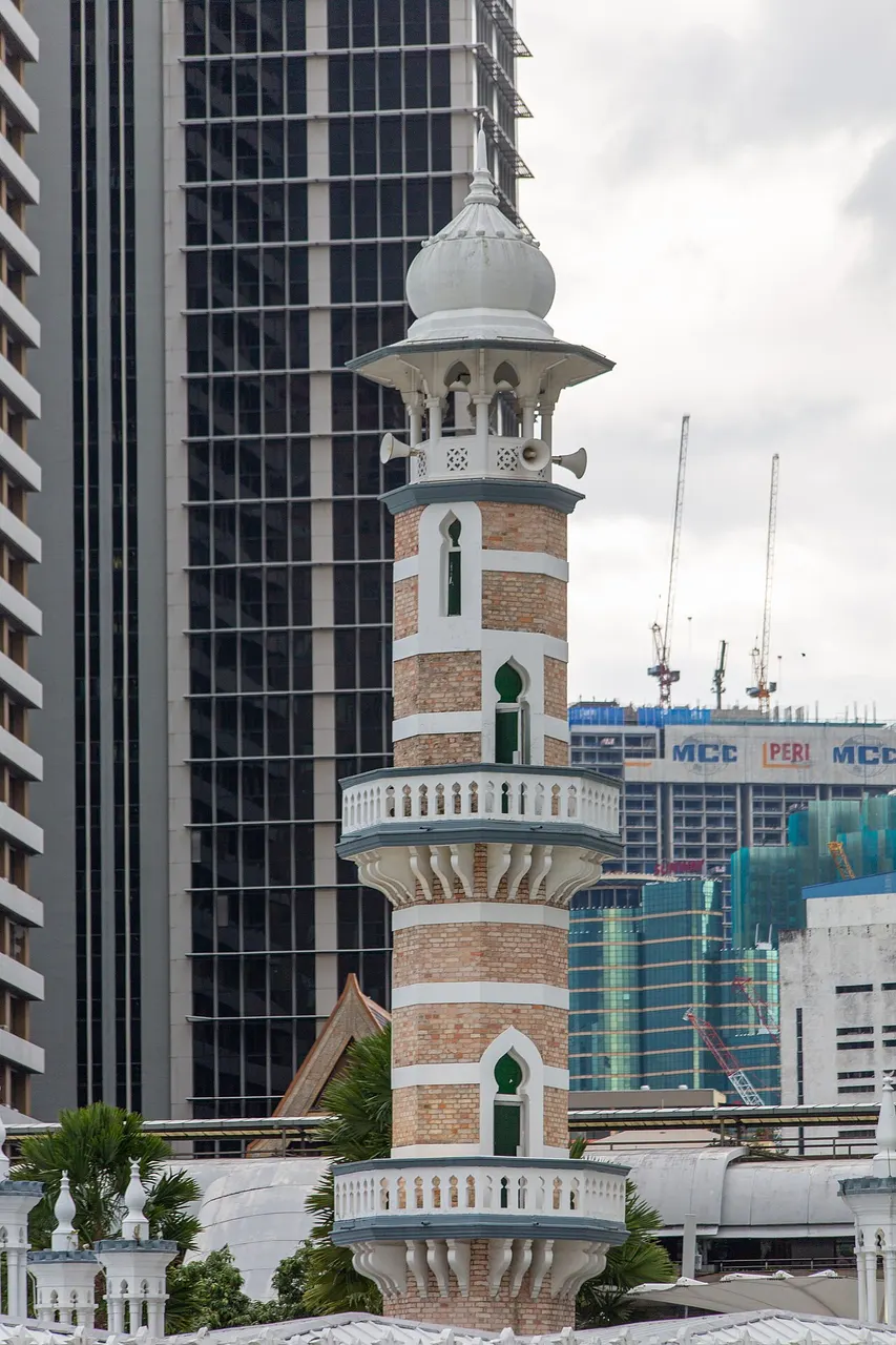 Minarete de faixas vermelhas e brancas com varandas e cúpula branca, entre prédios altos.