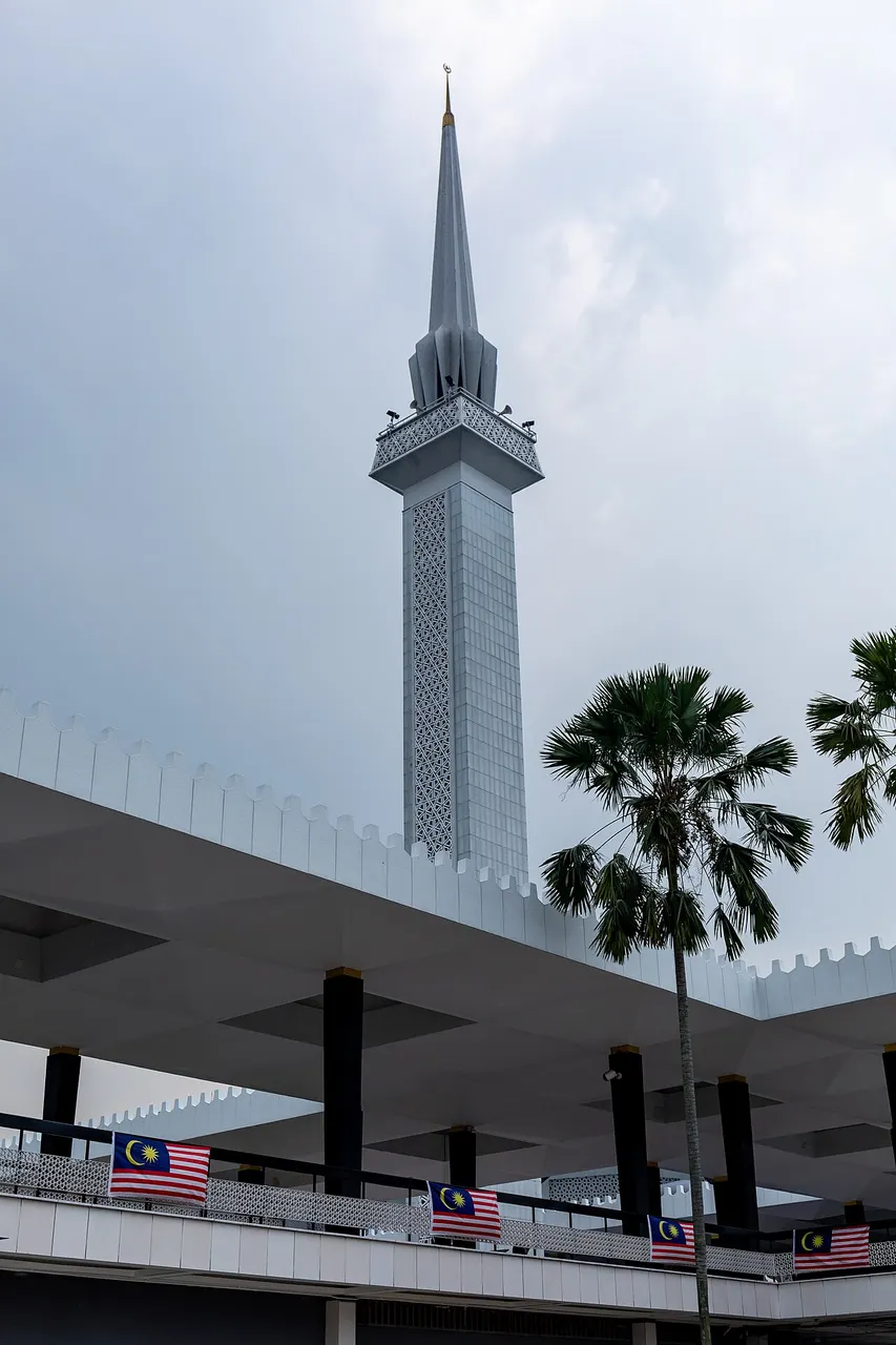Minarete moderno, alto e pontiagudo, de concreto claro, contra o céu nublado.