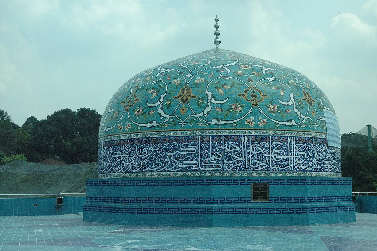 Cúpula de azulejos turquesa com faixa de caligrafia azul, sobre um tambor cilíndrico.