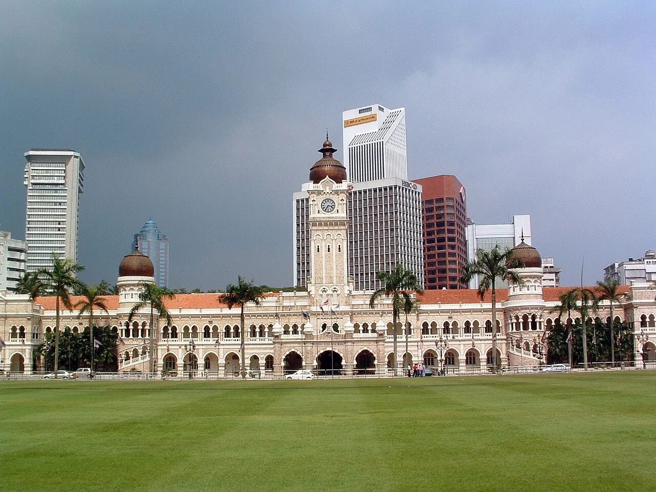 Edifício Sultan Abdul Samad visto do gramado, com a torre do relógio, cúpulas de cobre e palmeiras.