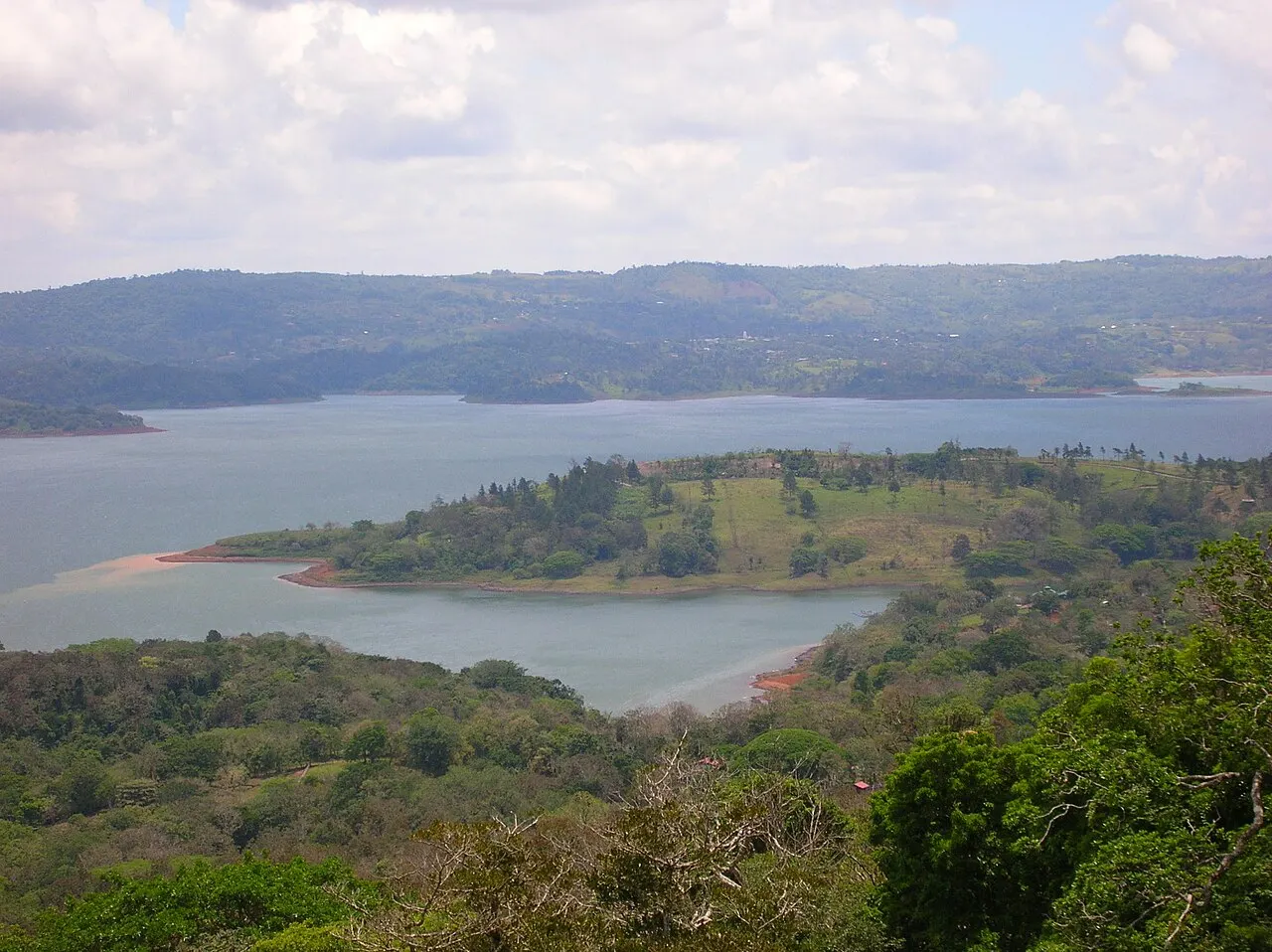 Vista do alto de um lago largo, com uma península de pasto e árvores avançando na água e morros baixos na outra margem.