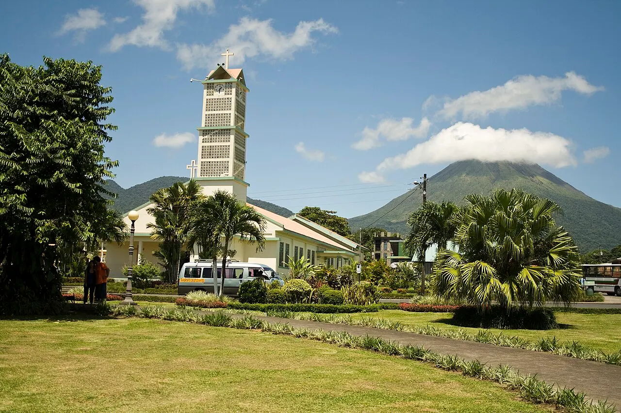 Um jardim com gramado, caminho e palmeiras diante de uma igreja de torre alta e quadrada, com o cone do vulcão parcialmente coberto de nuvens ao fundo.