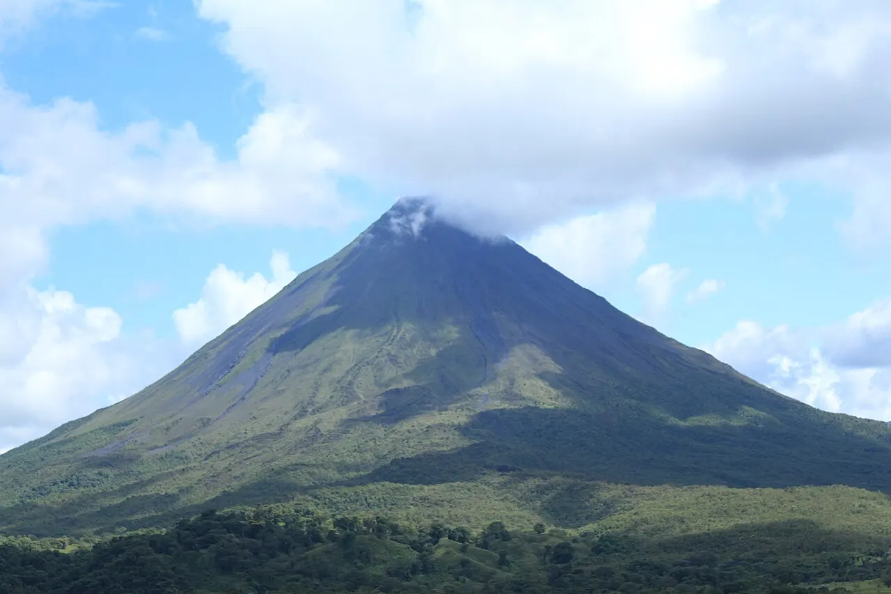 O cone quase simétrico do Volcán Arenal, coberto de verde nas encostas, com o cume escondido por uma nuvem e floresta na base.