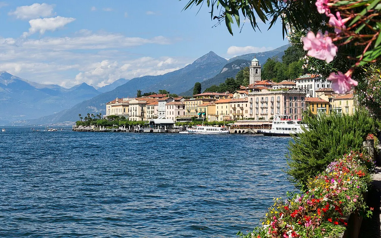 As casas coloridas de Bellagio na ponta do promontório, com barcos atracados, o lago azul e montanhas atrás, e flores em primeiro plano.