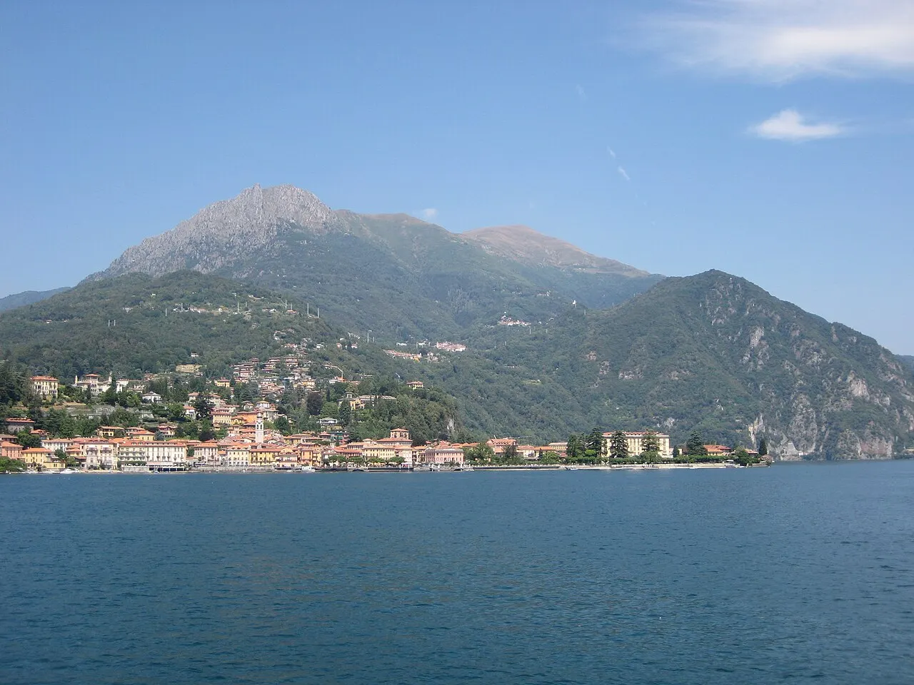 A cidade de Menaggio na margem do lago, vista da água, com casas claras na orla e montanhas atrás.