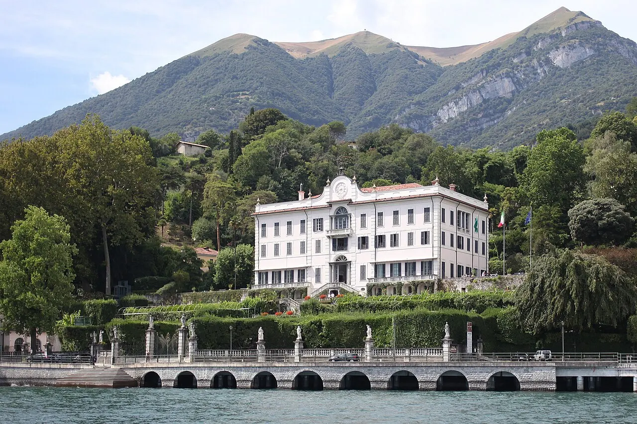 A Villa Carlotta branca vista do lago, com o jardim em degraus, a balaustrada do cais e as montanhas verdes atrás.