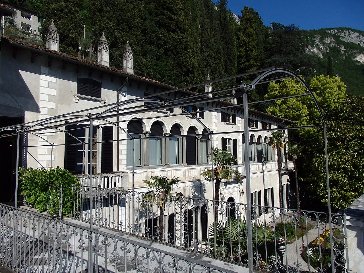 A fachada branca da Villa Monastero, com arcos no térreo, chaminés e uma pérgola de ferro no terraço sobre o jardim.