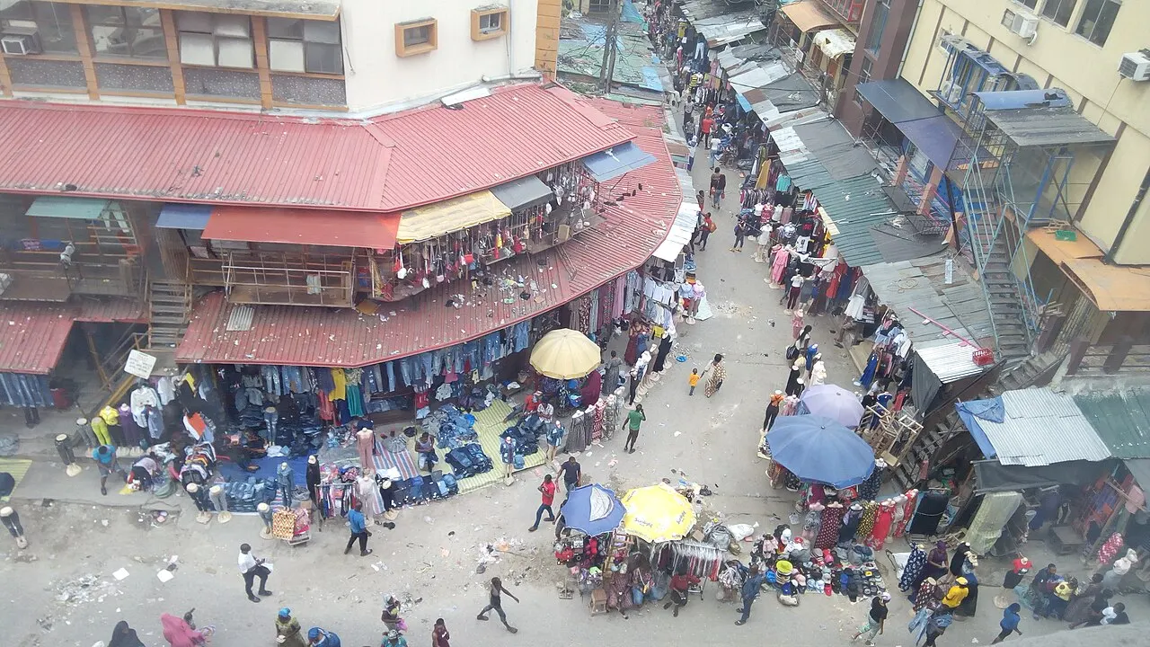 Vista aérea de ruas estreitas de mercado cobertas por telhados de zinco enferrujado, com barracas de roupas, guarda-sóis coloridos e pessoas circulando entre os corredores.