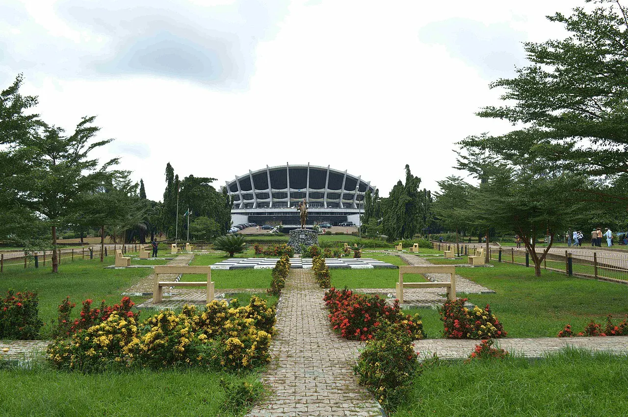 O volume curvo e nervurado do National Arts Theatre de Lagos ao fundo de um jardim, com caminho de pedra ao centro, canteiros de flores vermelhas e amarelas, bancos de concreto e uma estátua dourada sobre pedestal.