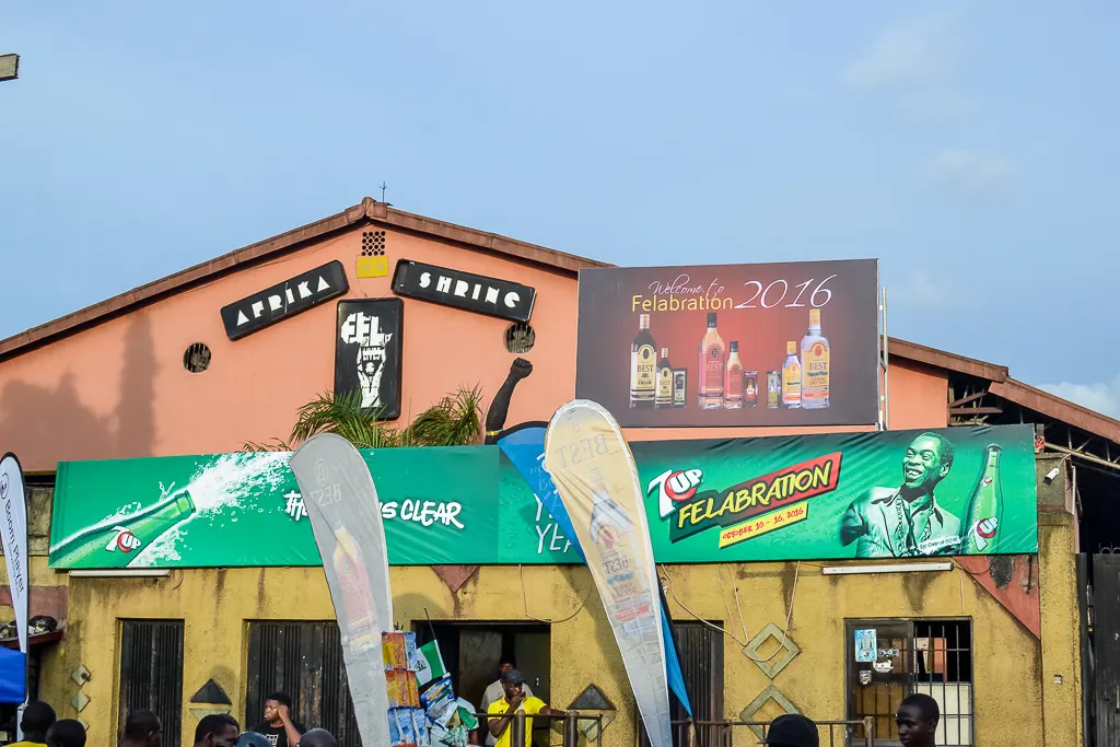 The facade of a building with a triangular roof, signs reading "Afrika Shrine", colorful advertising banners announcing a festival and people gathered at the entrance, under a blue sky.